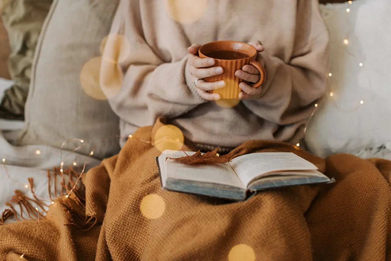 Person holding a warm drink in a cozy autumn setting with a blanket and book, illustrating November activities