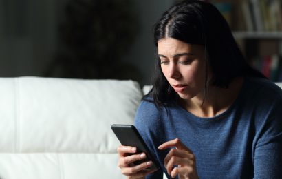 White woman sitting on a couch at night looking worried while reading a phone message
