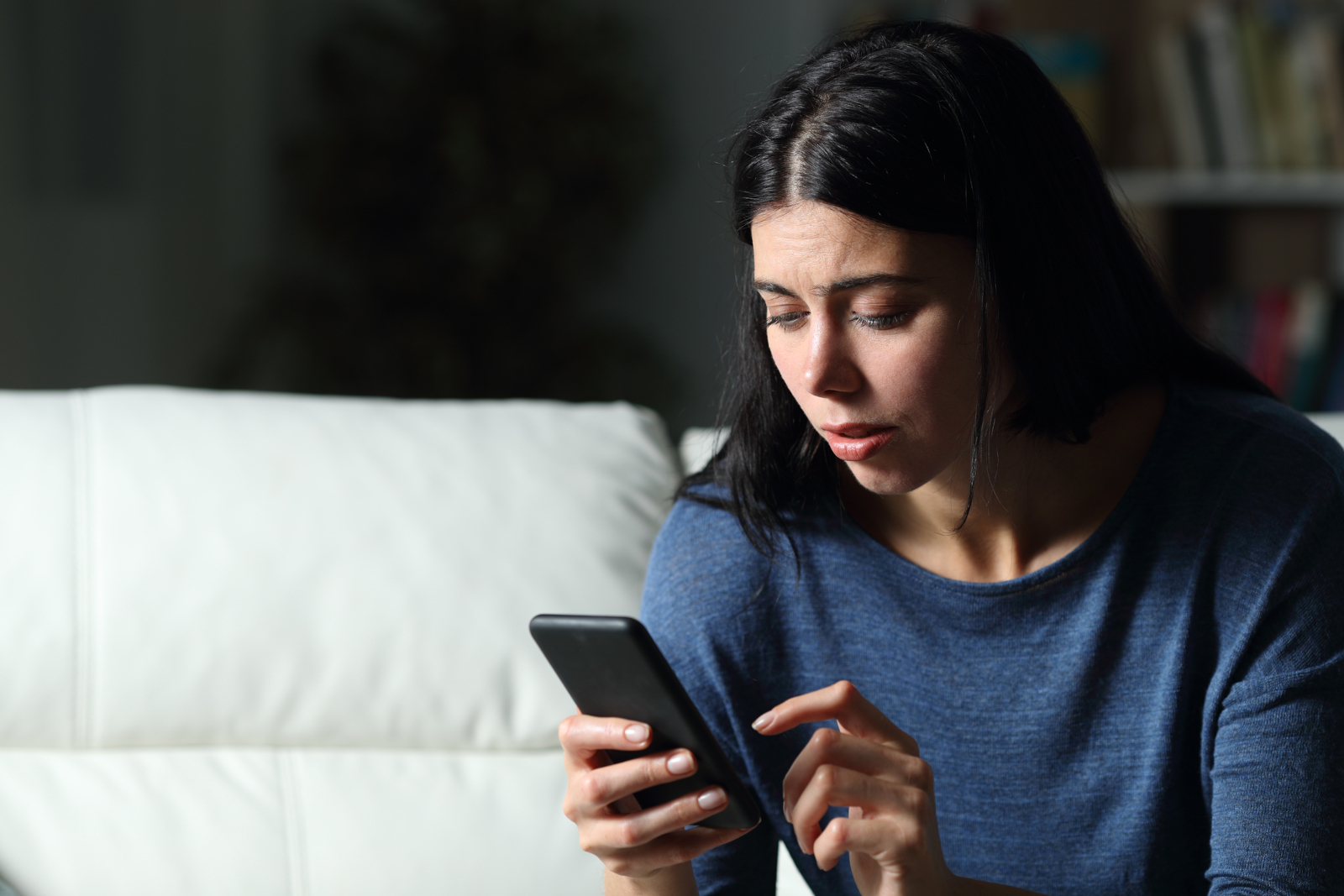White woman sitting on a couch at night looking worried while reading a phone message
