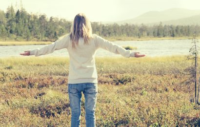 A woman from behind with arms open facing a scenic Nordic forest and lake landscape, practicing friluftsliv for stress relief.