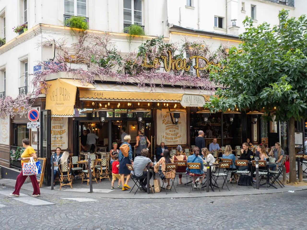 ibrant crowded outdoor terrace of Le Vrai Paris cafe in Montmartre with people dining under pink floral decorations highlighting authentic French lifestyle.