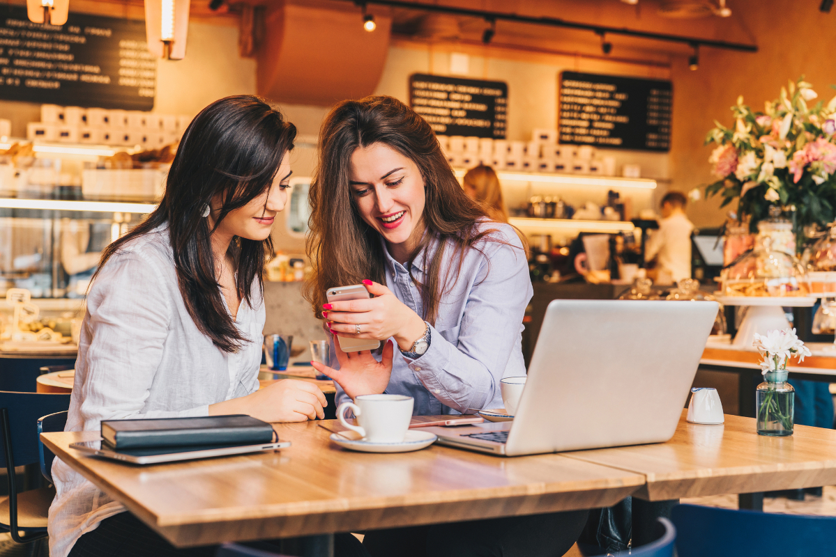Two women laughing at a cafe while using a smartphone and laptop