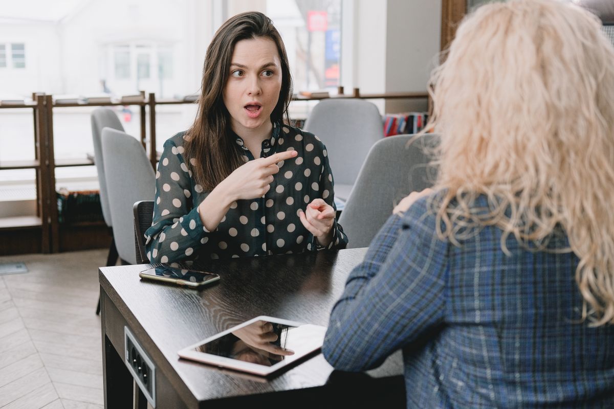 Two women having an intense conversation, with one looking shocked and pointing at the other.