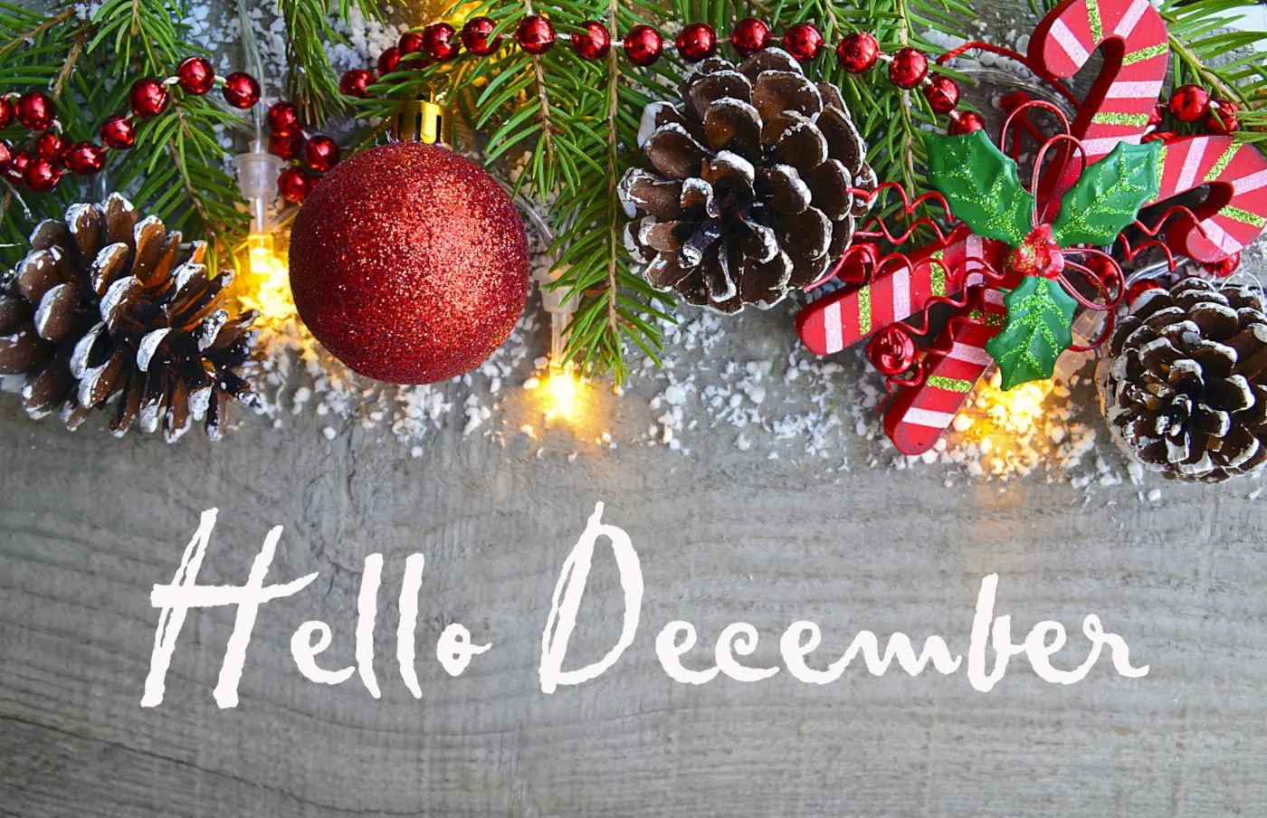 Hello December written on a wooden background decorated with evergreen branches, red Christmas ornaments, pinecones, and warm lights